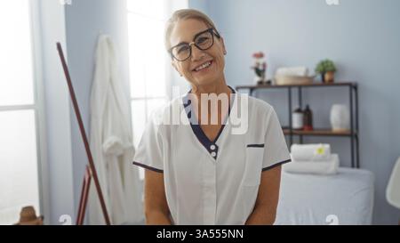 Donna sorridente in una stanza spa che indossa occhiali e uniforme bianca, incarna benessere, relax e cura professionale in un ambiente interno Foto Stock