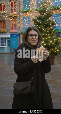 Donna che si gusta la pasticceria sulla strada di londra tra le festose luci natalizie nel cortile di neal, catturando il fascino delle vacanze invernali con decorazioni vivaci e joyfu Foto Stock