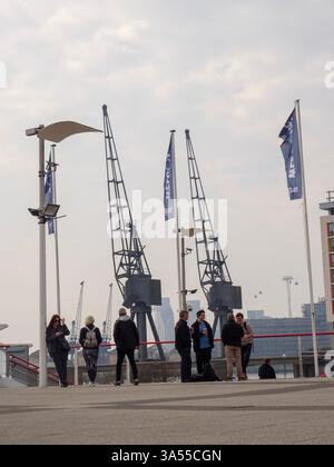 I partecipanti all'esposizione si fermano nell'atrio dell'Excel Centre all'esterno delle sale espositive con le storiche gru docklands sullo sfondo Foto Stock