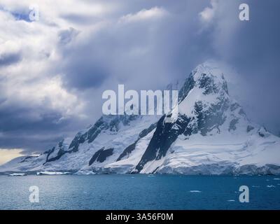 Una delle torreggianti montagne innevate che dominano l'Antartico con un bagliore di sole attraverso le nuvole che illuminano il mare. Foto Stock