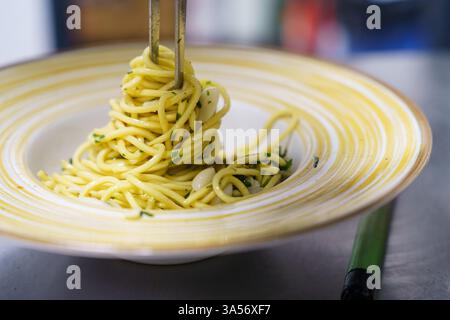 Uno shot ravvicinato di uno chef che toglie con cura spaghetti aglio e olio utilizzando pinze in una cucina professionale. La pasta è ricoperta di olio d'oliva, aglio, Foto Stock