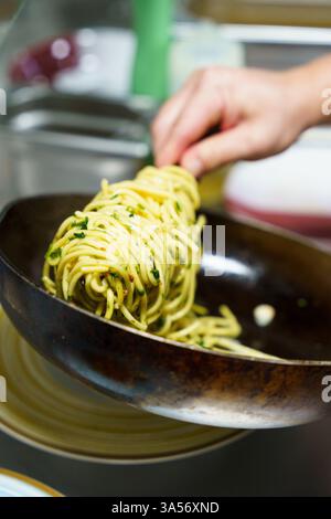 Uno chef prepara con cura una porzione di spaghetti aglio e olio utilizzando pinze in una cucina professionale. La pasta è ricoperta di olio d'oliva, aglio, prezzemolo, Foto Stock