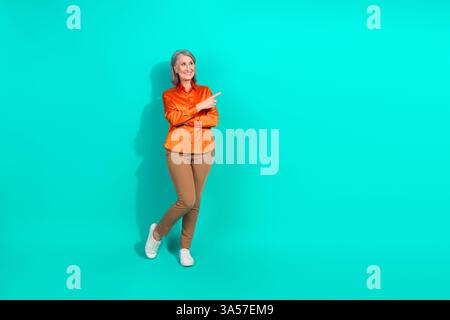 Donna matura che sorride con una vivace blusa arancione e pantaloni marroni che puntano da parte sullo sfondo turchese Foto Stock