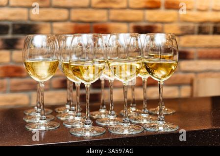 Una collezione di eleganti bicchieri da vino riempiti di vino bianco, disposti su un bancone del bar su uno sfondo rustico in mattoni. Foto Stock