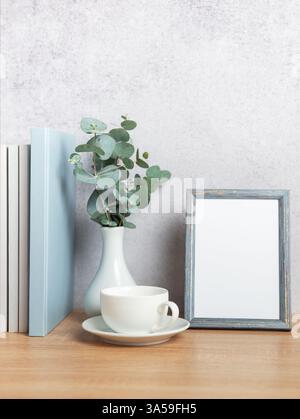 Interni dal design moderno con rami di eucalipto in vaso, cornice fotografica vuota, libri e tazza da caffè su tavolo in legno Foto Stock