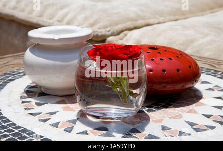 Tavolino da caffè con posacenere, candela e bicchiere con rosa Foto Stock