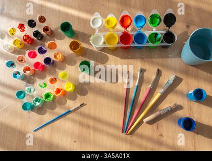 lattine aperte di pitture o gouache, vernici acriliche e pennelli si estendono su un tavolo in legno e illuminano i lati del sole Foto Stock