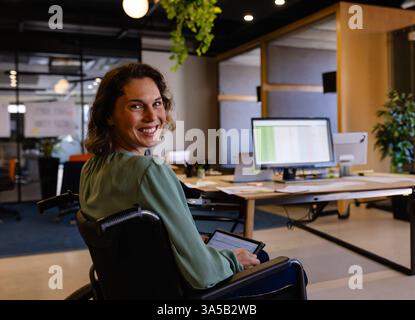Donna sorridente in sedia a rotelle che utilizza un tablet alla moderna scrivania dell'ufficio Foto Stock