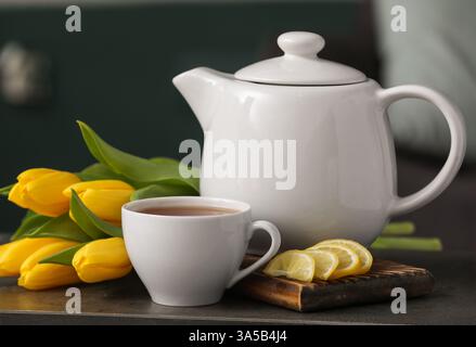 Tazza di tè caldo, fette di limone, pentola e tulipani sul tavolo su sfondo nero sfocato Foto Stock