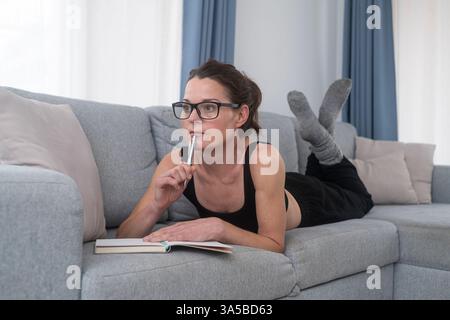 donna sdraiata su un divano che indossa occhiali da lettura con un taccuino e una penna Foto Stock
