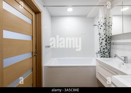 Bagno luminoso e pulito con piastrelle bianche, finiture moderne e una porta in legno con vetro smerigliato. Include una vasca da bagno con motivi geometrici Foto Stock