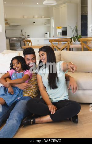 Famiglia felice seduta sul pavimento del soggiorno, genitori che puntano e figlia che sorride Foto Stock