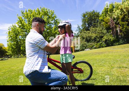 Padre che aiuta la figlia con il casco prima di andare in bicicletta nel parco soleggiato Foto Stock