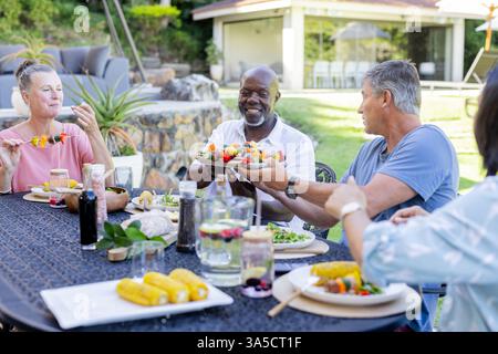 In giardino, diversi amici anziani condividono il cibo al barbecue, ridendo insieme all'aperto Foto Stock