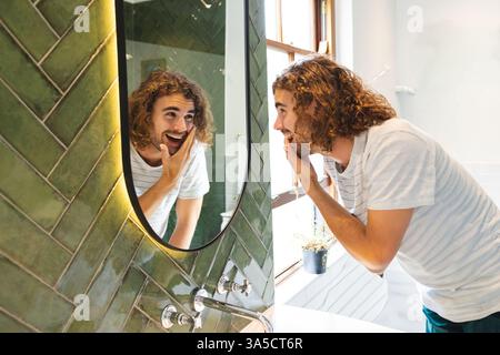 Giovane uomo con capelli ricci che ammira felicemente il riflesso nello specchio del bagno Foto Stock
