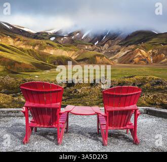 Viaggio in Islanda a luglio. Due chaise longue rosse collegate da un piccolo tavolo per i turisti. Montagne multicolore di riolite minerale sono l Foto Stock