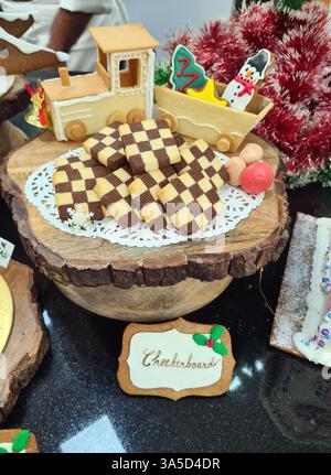 Biscotti a scacchi disposti ad arte su un piatto di legno, adornati con un treno giocoso e decorazioni festive per un'accogliente vetrina di panetteria Foto Stock