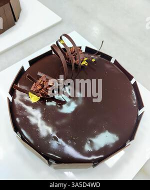 Una torta al cioccolato splendidamente lavorata adornata con ricci di cioccolato e accenti d'oro si trova in una panetteria, attirando i clienti. Foto Stock