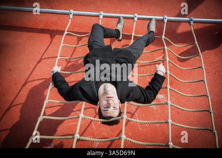 L'intera lunghezza di un serio uomo di 20 anni dai capelli scuri indossa una giacca nera e pantaloni sdraiati a testa in giù sulla corda di arrampicata, guardando intensamente la fotocamera Foto Stock