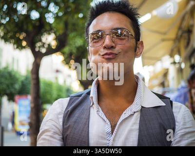 Giovane uomo etnico positivo in un abbigliamento casual e occhiali da vista alla moda sorridente con la bocca piena mentre mangia cibo in strada Foto Stock