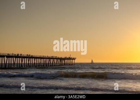 Una spiaggia di Hermosa al tramonto, caratterizzata da un lungo molo con un piccolo edificio e persone che camminano o pescano. Il cielo è un gradiente dal blu all'arancione Foto Stock