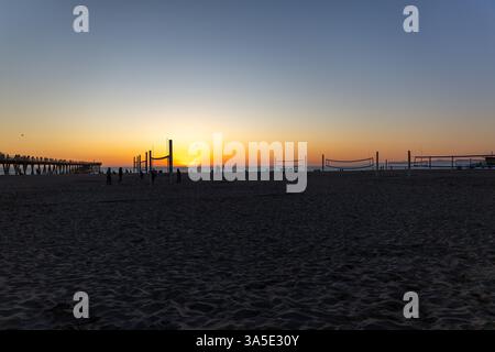 Una spiaggia di Hermosa al tramonto, caratterizzata da un lungo molo con un piccolo edificio e persone che camminano o pescano. Il cielo è un gradiente dal blu all'arancione Foto Stock