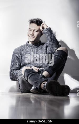 Bel giovane asiatico bianco che indossa un maglione grigio e jeans strappati, seduto su uno sfondo chiaro contro le pareti in studio Foto Stock