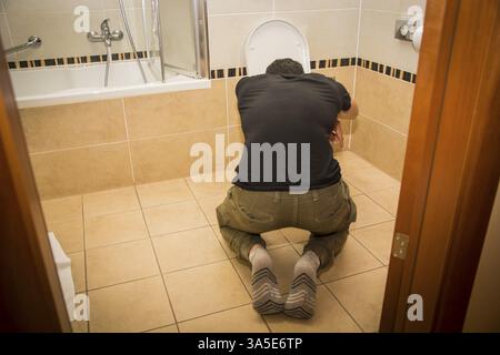 Vista posteriore di un giovane ubriaco che vomita nella toilette a casa mentre è in posizione di ginocchio Foto Stock