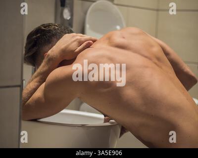 Vista posteriore di un giovane ubriaco che vomita nella toilette a casa mentre è in posizione di ginocchio Foto Stock