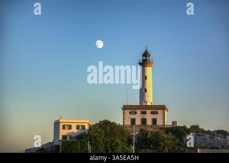 Vista panoramica di un faro sulla cima di una collina al tramonto, con un bagliore arancione nel cielo e sulla luna per avvisare le navi di un pericolo di navigazione e fornire Foto Stock