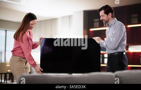 Marito e moglie sorridenti spostano con cura la nuova televisione nell'appartamento. Foto Stock
