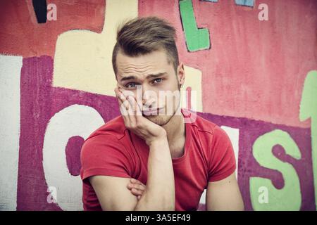 Bella bionda giovane uomo contro il coloratissimo graffito parete facendo espressione carino Foto Stock