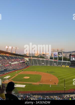Vista grandangolare di una partita di baseball in un grande stadio durante il tramonto, con giocatori sul campo e spettatori in tribuna sullo sfondo della città. Foto Stock