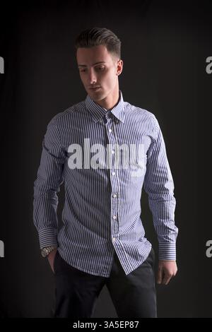 Giovane sorridente in camicia a righe, con lo sguardo rivolto verso il basso su sfondo scuro Foto Stock