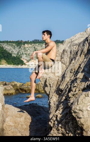 Attraente bel giovane uomo seduto sulla roccia dal mare o ocean shore con capelli bagnati, guardando in lontananza, vista di profilo Foto Stock