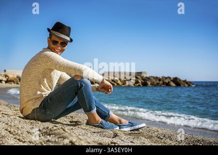 Foto a tutta lunghezza di un bel giovane atletico in abbigliamento trendy, in spiaggia in un giorno d'estate soleggiato, guardando la fotocamera sorridente, contro il Blue Sky dietro Foto Stock