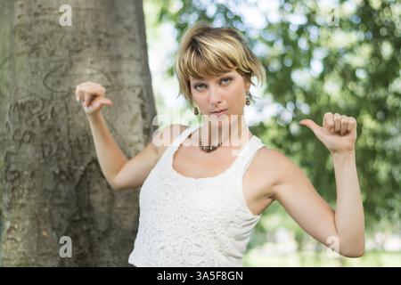 Attraente giovane donna bionda all'aperto che punta le dita verso se stessa, sicura di sé mentre guarda la fotocamera Foto Stock
