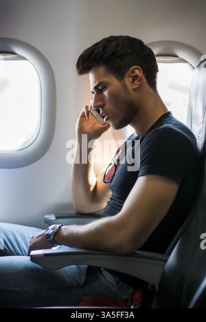 Vista laterale del bel giovane uomo contro la finestra dell'aereo seduto e che parla al telefono Foto Stock