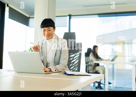 In un ufficio chic, una donna asiatica concentrata con un abito su misura si adatta al suo laptop, regolando gli occhiali mentre un collega lavora nelle vicinanze, mostrando prodotti Foto Stock