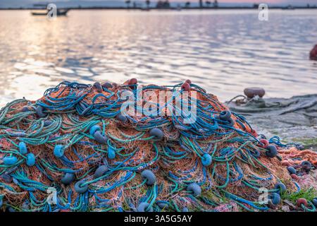Reti da pesca con corde blu e verdi e galleggianti rossi accumulati vicino a un mare calmo al tramonto, catturati in luce soffusa. Foto Stock