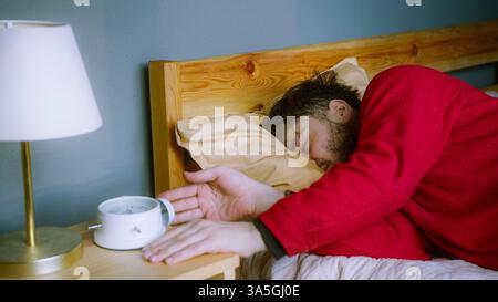 Il giovane lottava per svegliarsi, colpendo Snooze alla sveglia Foto Stock