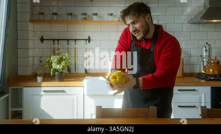 Giovane uomo in una cucina accogliente che tiene in mano una fresca Quince gialla, indossa un grembiule nero e sorride mentre prepara un pasto Foto Stock