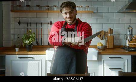 Uomo sicuro di sé in una cucina moderna con un coltello affilato, un grembiule nero e un maglione rosso, sorridendo con un atteggiamento professionale da chef Foto Stock