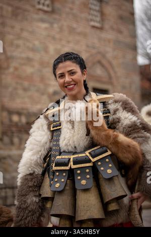 Kyustendil, Bulgaria – 15 febbraio 2025: Il "Djamala" Masquerade Festival. Una donna in pelle di pecora e una sciarpa rossa con un volto dipinto. Dietro di lei, una W Foto Stock