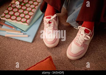Eleganti sneakers indossate da una persona seduta su pavimento tappezzato con libri, matite e notebook nelle vicinanze, creando una scena accogliente in un ambiente informale Foto Stock