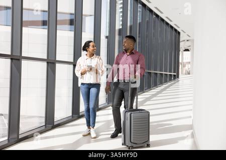 Due giovani colleghi viaggiano insieme per lavoro, camminano attraverso un corridoio moderno, fanno rotolare le valigie e parlano. La coppia arriva all'aeroporto e chiama Foto Stock
