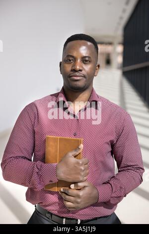 Ritratto verticale di un uomo d'affari, CEO o membro del personale aziendale africano professionista, vestito con una camicia casual che tiene in mano l'organizzatore personale, sta Foto Stock