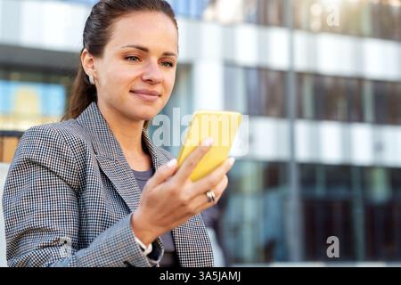 Donna d'affari sicura e sorridente che utilizza lo smartphone all'aperto per le strade della città Foto Stock