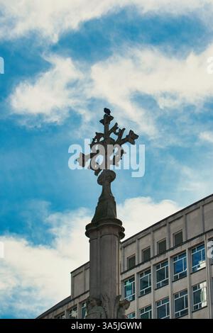 Monumento in pietra con croce ornata sormontata da un uccello, adagiato su un cielo blu con nuvole e un edificio moderno a Manchester, Regno Unito. Foto Stock