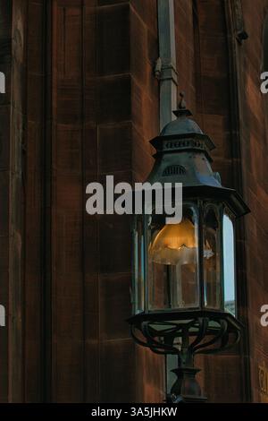 Una lanterna ornata è appesa al muro di un edificio, che emette un caldo bagliore. L'edificio ha una facciata in pietra con un design verticale a Manchester, Regno Unito. Foto Stock